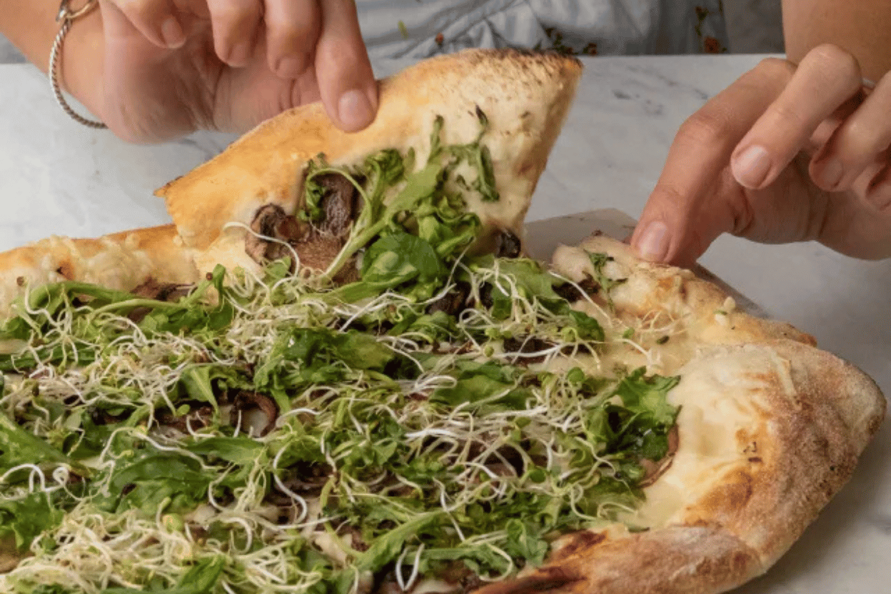 Woman lifting a slice of pizza topped with mushrooms, arugula, and sprouts