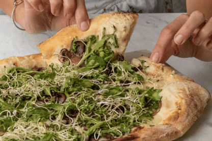 Woman lifting a slice of pizza topped with mushrooms, arugula, and sprouts